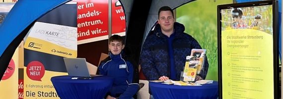 2 Männer sitzen an blauem Infostand an einem Tisch und lächeln